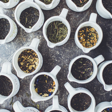 White tea cups filled with various types of loose leaf tea on a dark surface.