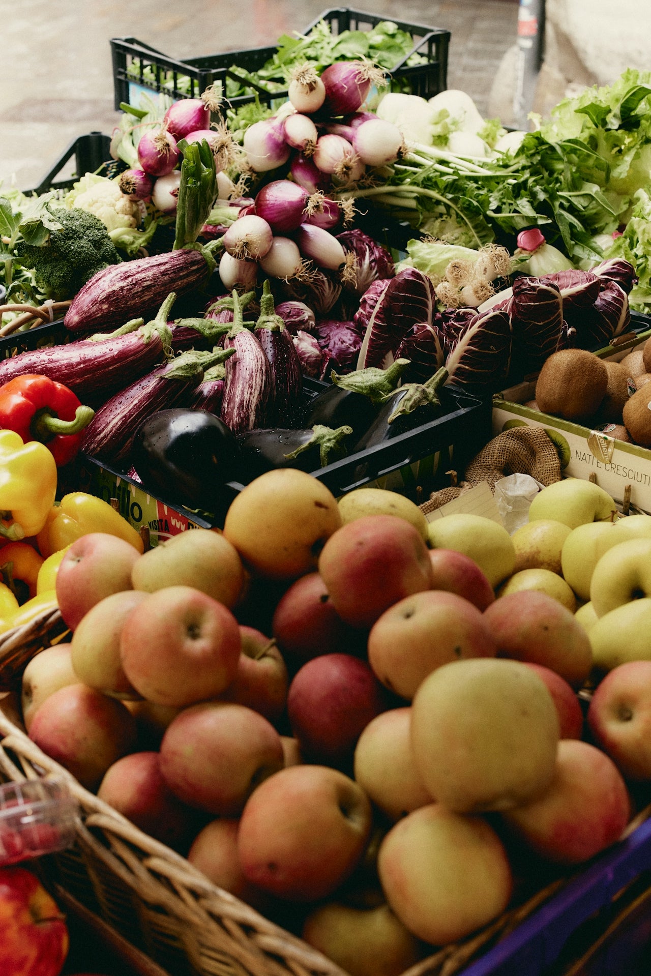 Assorted fruits and vegetables including apples, eggplants, and leafy greens at a market stall.